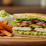 Chicken Avocado Sandwich with grilled chicken, creamy avocado, fresh vegetables, and toasted sourdough bread, served with sweet potato fries.