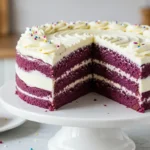 A moist and fluffy Purple Velvet Cake with vibrant purple layers, topped with smooth cream cheese frosting, elegantly displayed on a white cake stand.