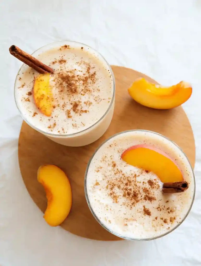 Creamy Peach Smoothie (No Sugar Needed) 6 Two peach smoothies topped with cinnamon and peach slices on a round wooden board