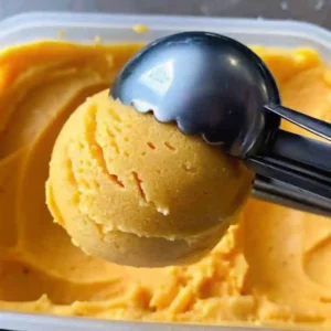 A metal scoop lifting a round scoop of mango ice cream from a freezer container