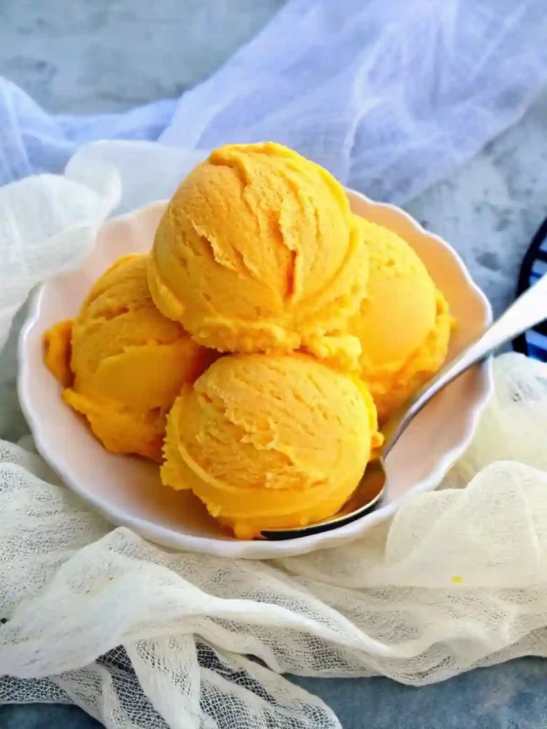 Homemade Mango Ice Cream (No-Cook, No-Egg) 4 Four scoops of mango ice cream in a white bowl with a spoon and light fabric background
