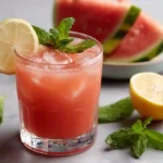 A chilled glass of watermelon lemonade garnished with a lemon slice and mint, with watermelon and lemons in the background.