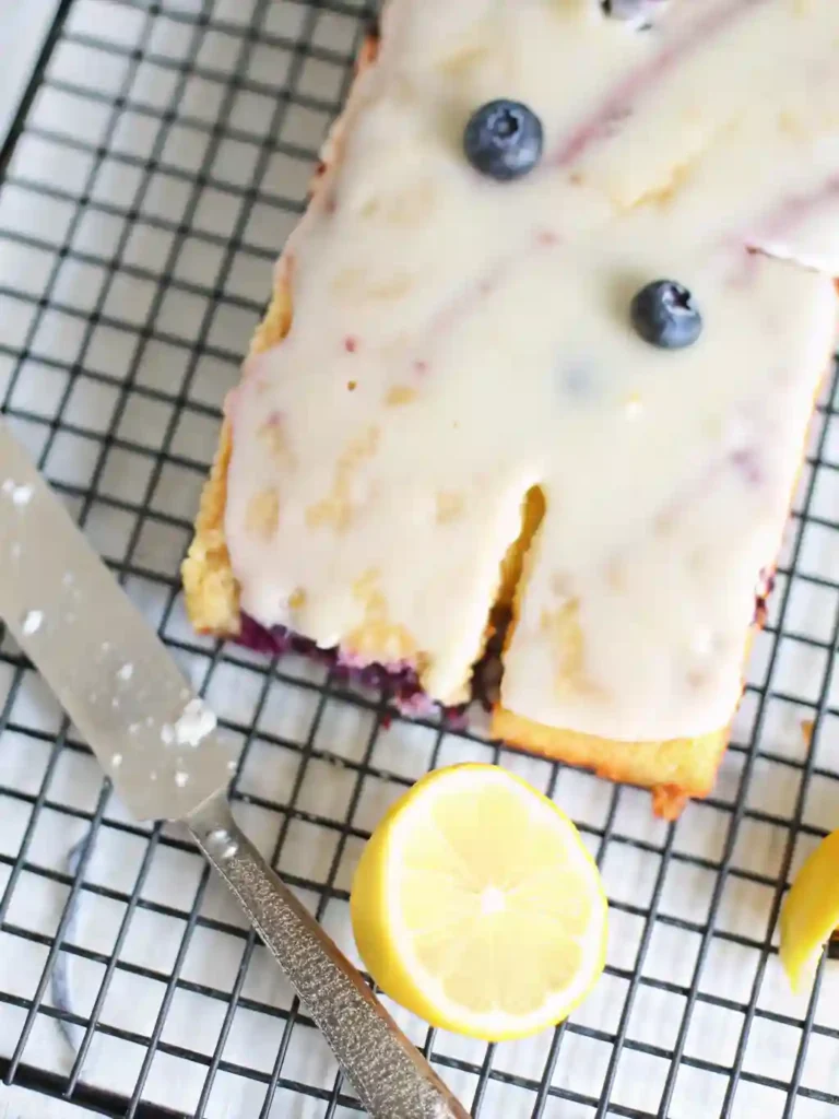 Lemon Blueberry Sheet Cake Recipe 4 Sliced lemon blueberry sheet cake with lemon glaze and blueberries on top, shown on a wire rack with lemon halves