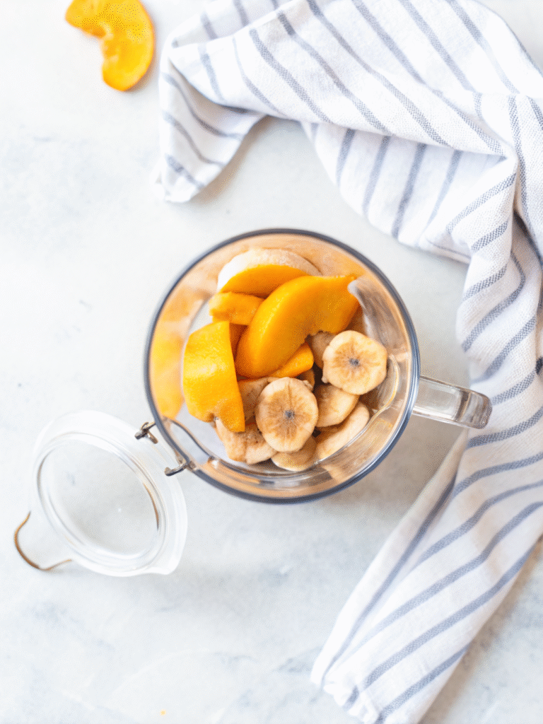Creamy Peach Smoothie (No Sugar Needed) 3 Sliced peaches and bananas in a blender cup on marble with striped towel