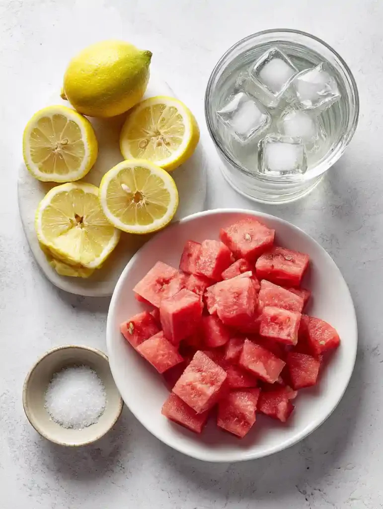 Fresh Watermelon Lemonade: The Perfect 5-Ingredient Summer Refresher 2 Flat lay of chopped watermelon, sliced lemons, a glass of ice water, and a small bowl of sugar on a light surface.
