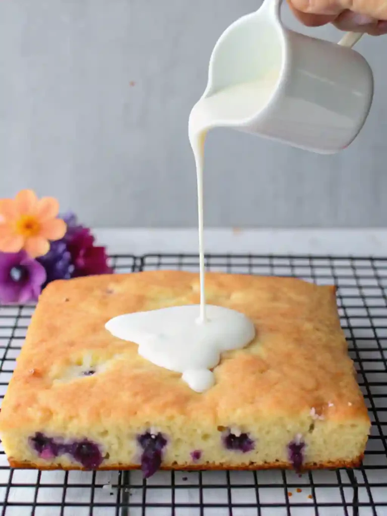 Lemon Blueberry Sheet Cake Recipe 3 Lemon blueberry sheet cake on a wire rack with lemon glaze being poured from a white pitcher