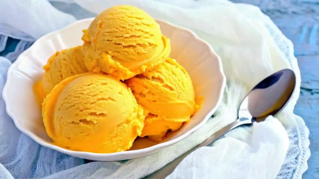 Close-up of mango ice cream scoops served in a white bowl next to a spoon on a soft cloth background