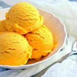 Homemade Mango Ice Cream (No-Cook, No-Egg) 12 Close-up of mango ice cream scoops served in a white bowl next to a spoon on a soft cloth background