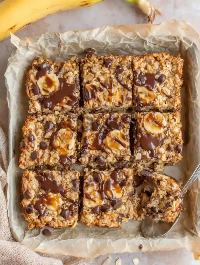 Banana Oatmeal Bars Recipe | Healthy Breakfast Snack 2 Freshly baked banana oatmeal bars with chocolate drizzle and banana slices in a parchment-lined pan