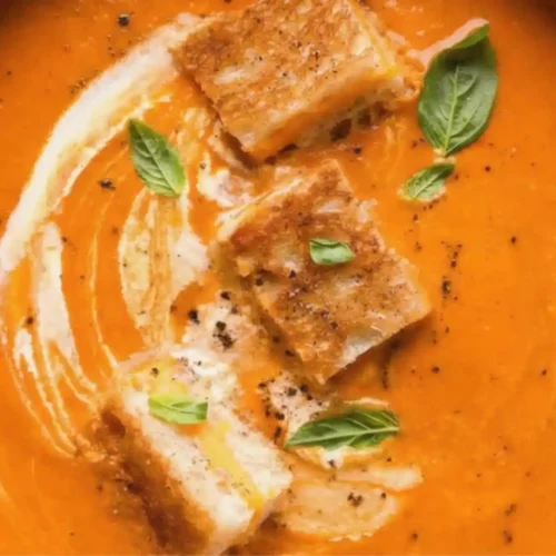 Bowl of Homemade Tomato Soup topped with cream swirl, basil leaves, and grilled cheese croutons