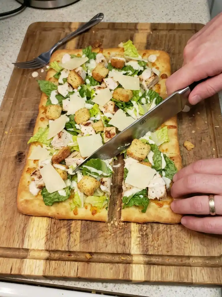 Chicken Caesar Flatbread being sliced on a wooden board with grilled chicken, romaine, Parmesan, and Caesar drizzle