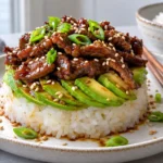 Close view of a teriyaki beef and avocado rice stack with glossy sliced beef fanned avocado and sticky rice garnished with sesame seeds and green onions with chopsticks on the plate